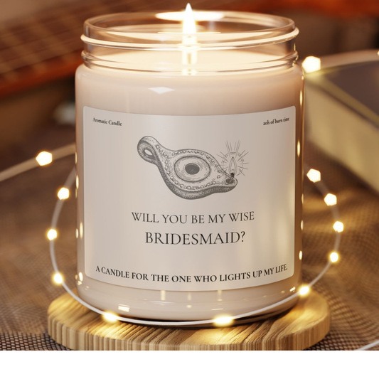 Candle in a jar with a label asking 'Will you be my wise bridesmaid?' on a wooden coaster with string lights.
